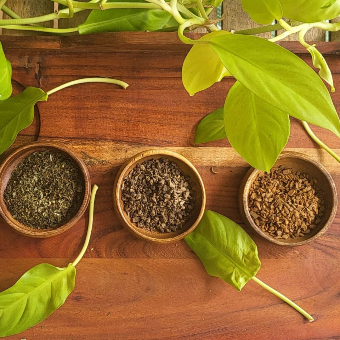 Three wooden bowls with dried herbs on a wooden surface with green leaves.