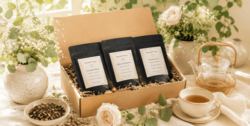 A kraft box filled with kraft paper and 3 packages in the box placed beautifully. The box is surrounded with herbs, flowers in a vase and a ceramic tea kettle.