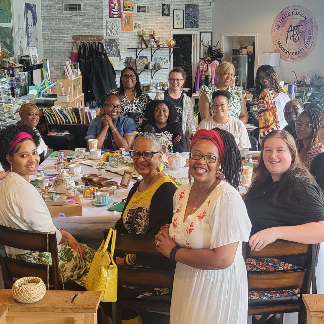 A group of women gathered around a table in a creative studio or workshop, smiling and posing together. The room has a white brick wall, shelves, and various items in the background.