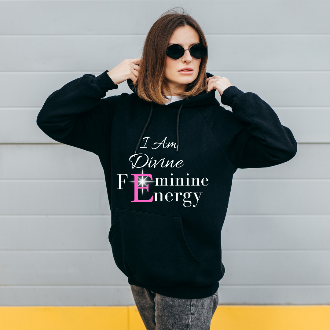 Woman wearing a black hoodie with 'I Am Divine Feminine Energy' text against a gray wall.