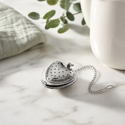 Heart-shaped tea infuser on a marble surface with a cup and greenery in the background