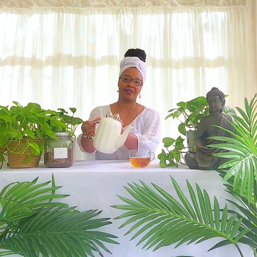 A woman wearing a white headwrap and glasses pours tea from a white teapot into a glass cup on a white tablecloth-covered table. The scene is decorated with potted green plants, a glass jar, and a Buddha statue, set against a background of sheer white curtains.