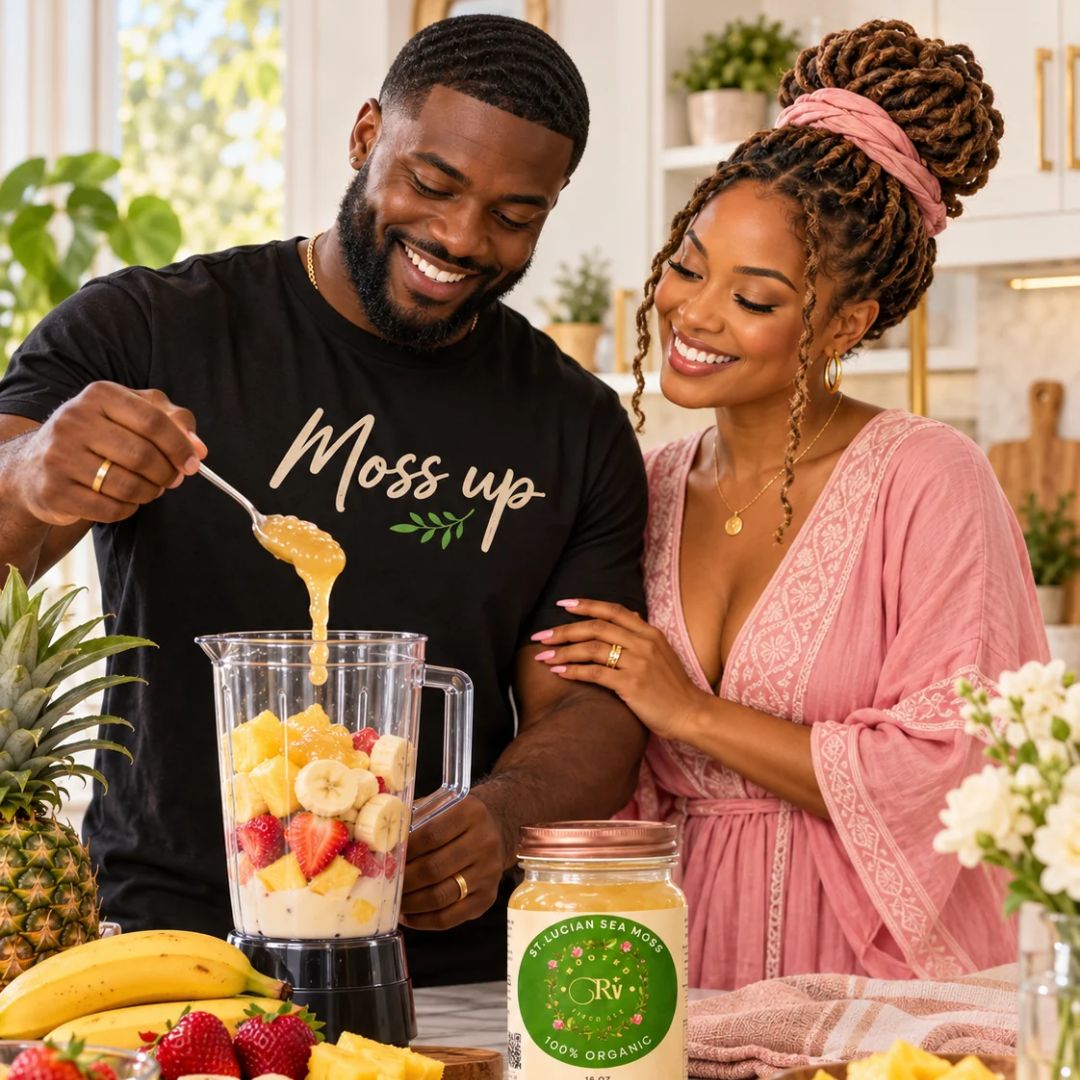 A man and a woman smile while preparing a smoothie in a bright kitchen. The man wears a black t-shirt with 'Moss up' printed on it, and the woman wears a pink robe.