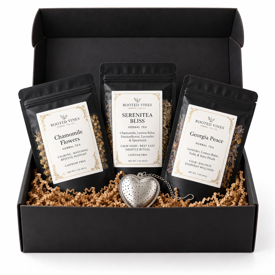 A black gift box filled with brown crinkle paper shred, holding three black herbal tea pouches from Rooted Vines: Chamomile Flowers, Serenitéa Bliss, and Georgia Pencil, with a silver heart-shaped keychain in front.