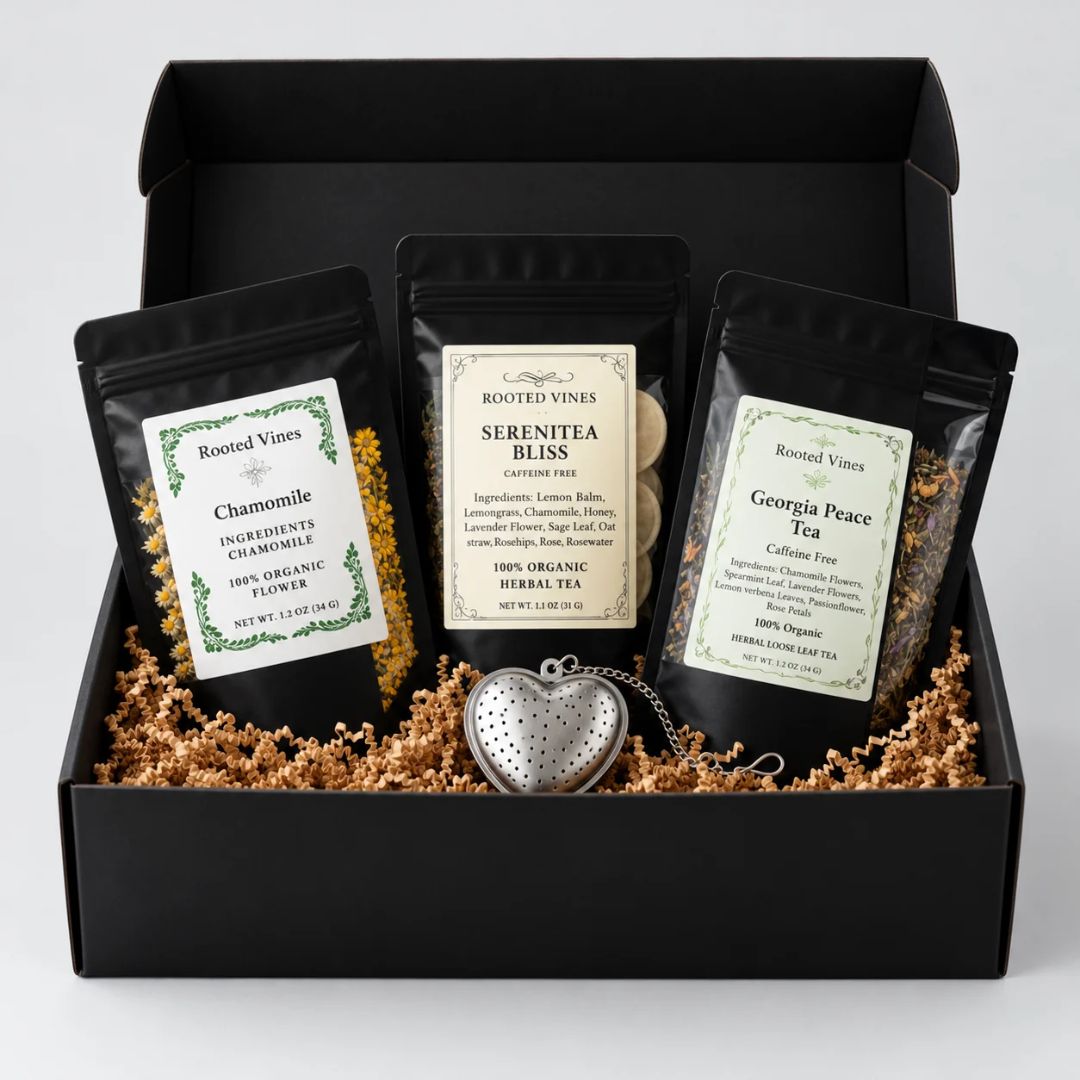 A black gift box filled with shredded brown paper filler holds three black tea bags with cream labels and a small silver heart-shaped metal tea infuser. The labels read Rooted Vines and show Chamomile, Serenitea Bliss, and Georgia Peace Tea.