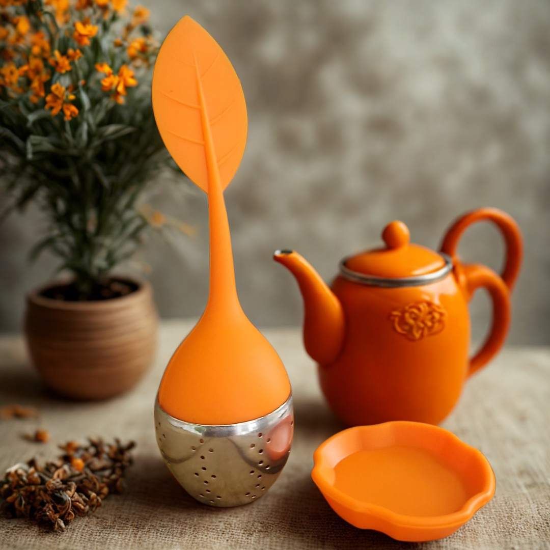 Orange tea infuser, teapot, and leaf-shaped spoon on a textured surface with a plant in the background.