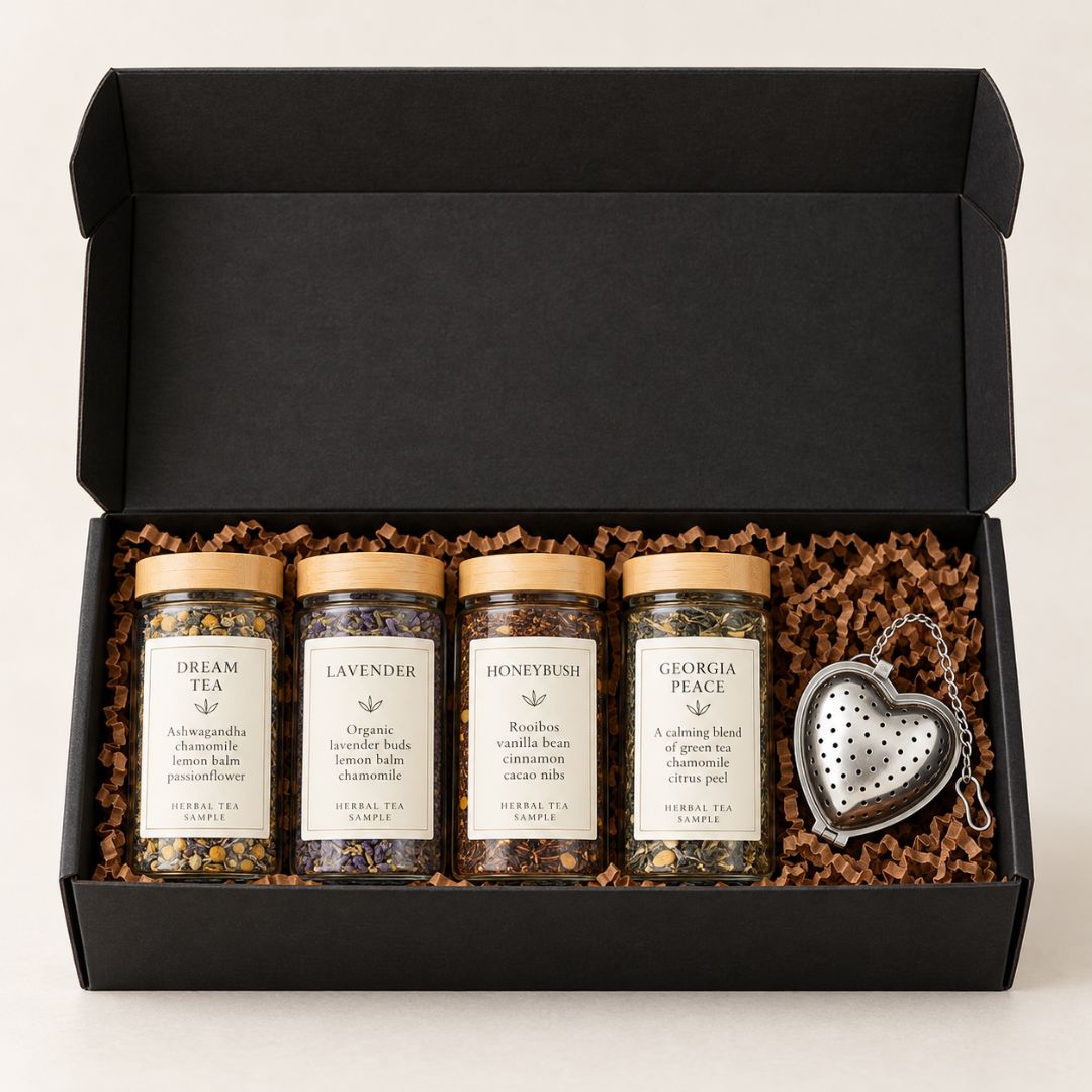 A black gift box containing four glass jars of herbal tea with gold lids and white labels reading Dream Tea, Lavender, Honeybun, and Georgia Place, arranged in brown crinkle paper with a silver heart-shaped metal tea infuser.