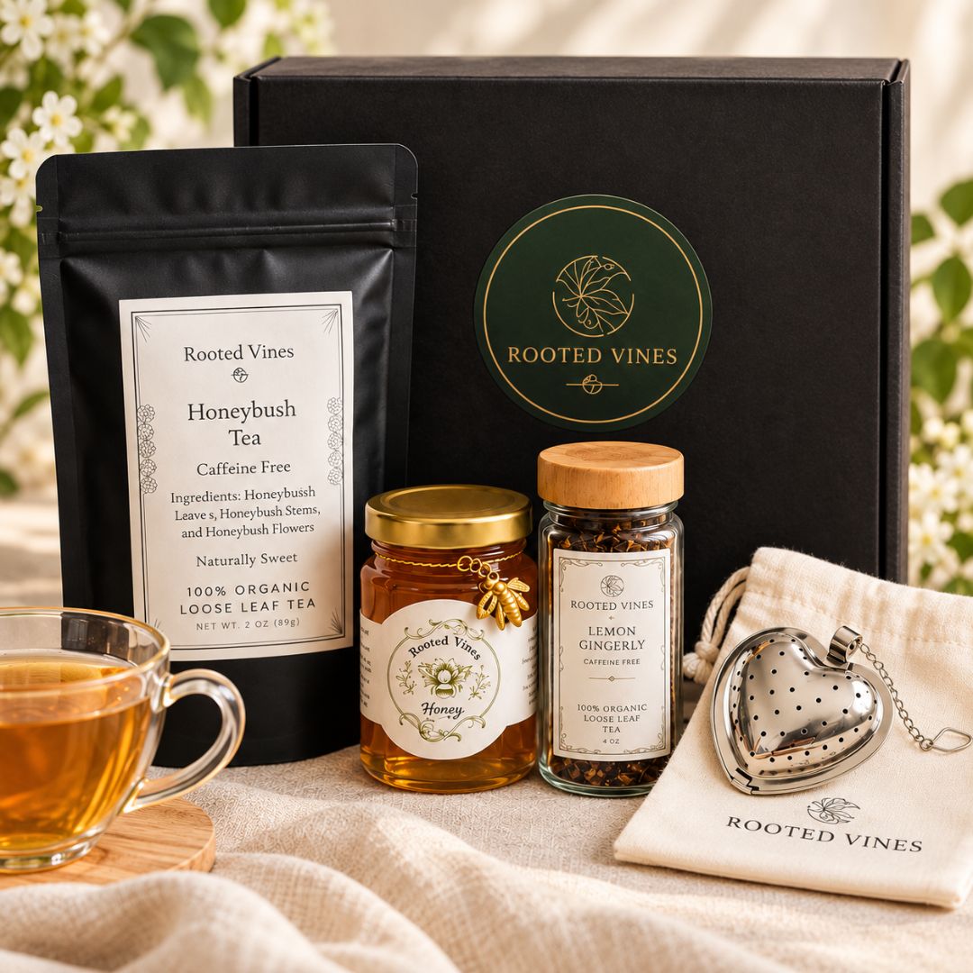 Rooted Vines tea gift set featuring a black pouch of honeybush tea, a jar of lemon gingerly, a glass jar of loose leaf tea, a heart-shaped silver teapot on a cream cloth, and a black gift box with a green circular logo, arranged on a beige fabric surface with a glass of tea in the foreground.